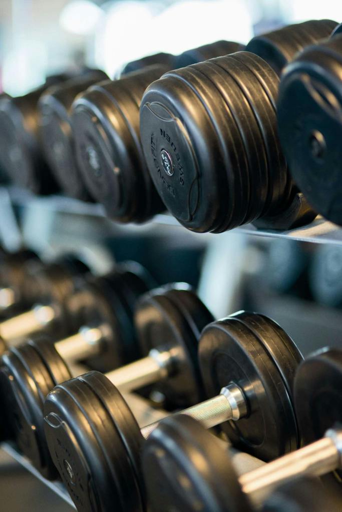Close-up of black dumbbells arranged in a weight rack at a gym. Multiple dumbbells with metal handles are visible in rows, showing various weight sizes with black plates. The image has a shallow depth of field, creating a professional fitness equipment aesthetic.